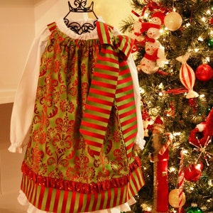 Puede incluir: Un vestido de Navidad verde y rojo con un lazo a rayas rojo y verde. El vestido tiene un dobladillo con volantes y está sobre un soporte blanco delante de un árbol de Navidad decorado.