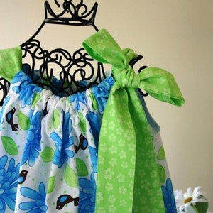 May include: A sleeveless dress with a floral pattern in blue and white, accented with green leaves and small birds. The dress features green bows on the shoulders. The dress is displayed on a black metal form.