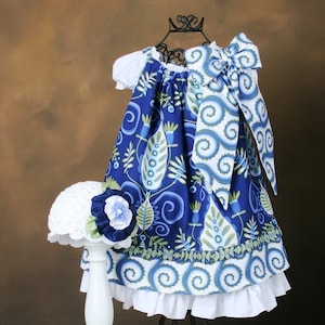 May include: A blue and white floral print dress with a white ruffle trim and a matching white hat. The dress has a bow at the neckline and is displayed on a black stand.