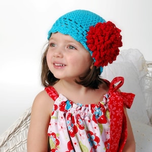May include: A young girl wearing a turquoise crocheted hat with a red crocheted flower on top. She is wearing a white dress with red and blue floral print.