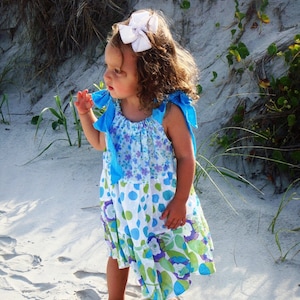 Puede incluir: Una ni&ntilde;a peque&ntilde;a con un lazo blanco en el pelo y un vestido floral azul y blanco con lunares. Est&aacute; de pie en una playa de arena.