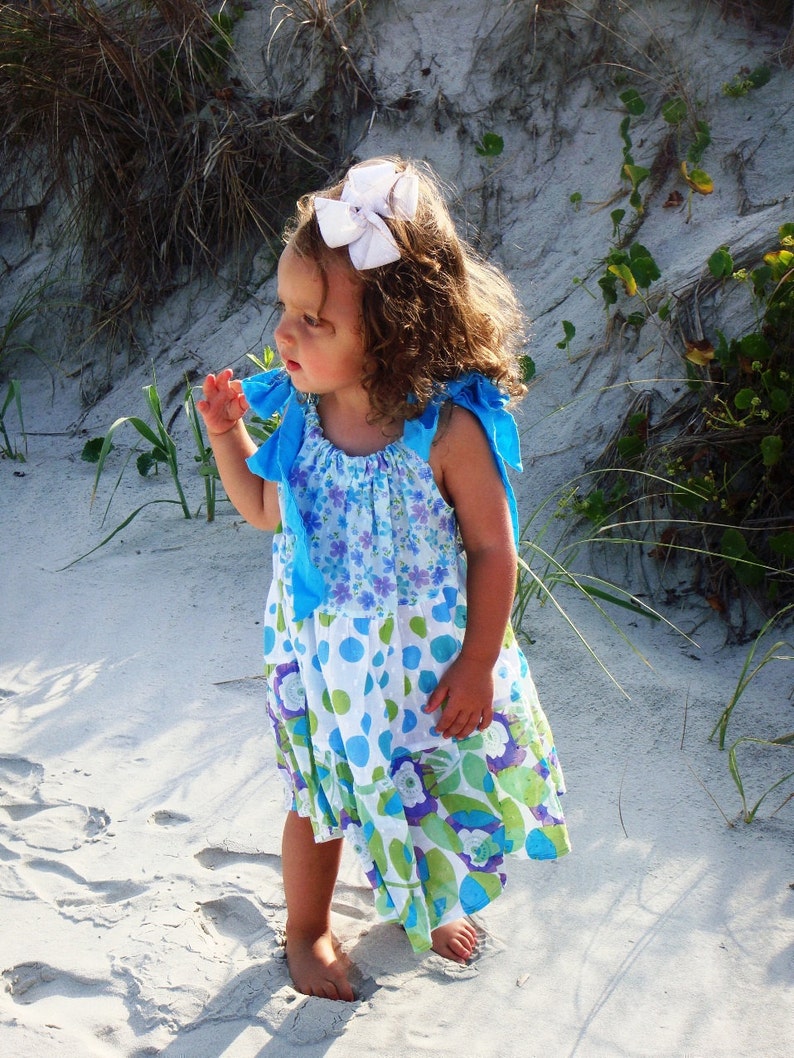 Puede incluir: Una ni&ntilde;a peque&ntilde;a con un lazo blanco en el pelo y un vestido floral azul y blanco con lunares. Est&aacute; de pie en una playa de arena.