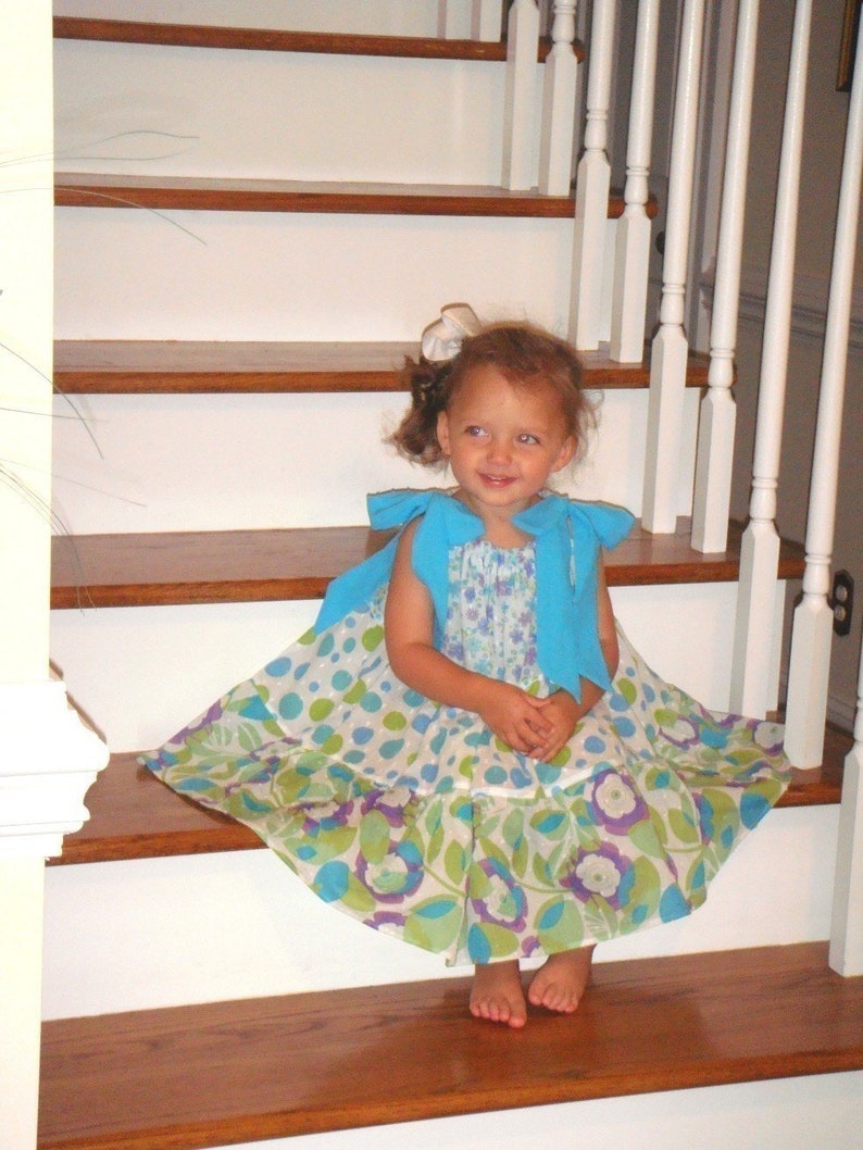 May include: A young girl wearing a blue and white floral dress with a blue cardigan sits on a wooden staircase.