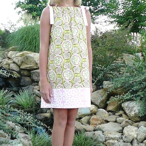 Puede incluir: Una niña pequeña con un vestido de estampado floral verde y rosa con tirantes blancos. El vestido tiene un dobladillo de estampado floral rosa y blanco. Está de pie sobre una roca grande con un fondo rocoso.