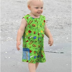 May include: A young child wearing a sleeveless green romper with a colorful pattern of tools and objects. The romper has a button-up front and a patch of blue and white stripes. The child is walking on a sandy beach.