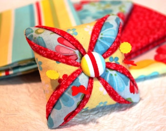 May include: A yellow, blue, and red pincushion with a red, white, and yellow button in the center. The pincushion is made of fabric with a floral pattern and has several pins sticking out of it.