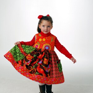 Puede incluir: Una niña pequeña con un vestido de retazos rojo y multicolor, un cuello alto rojo y un lazo rojo en el pelo. Lleva botas marrones.