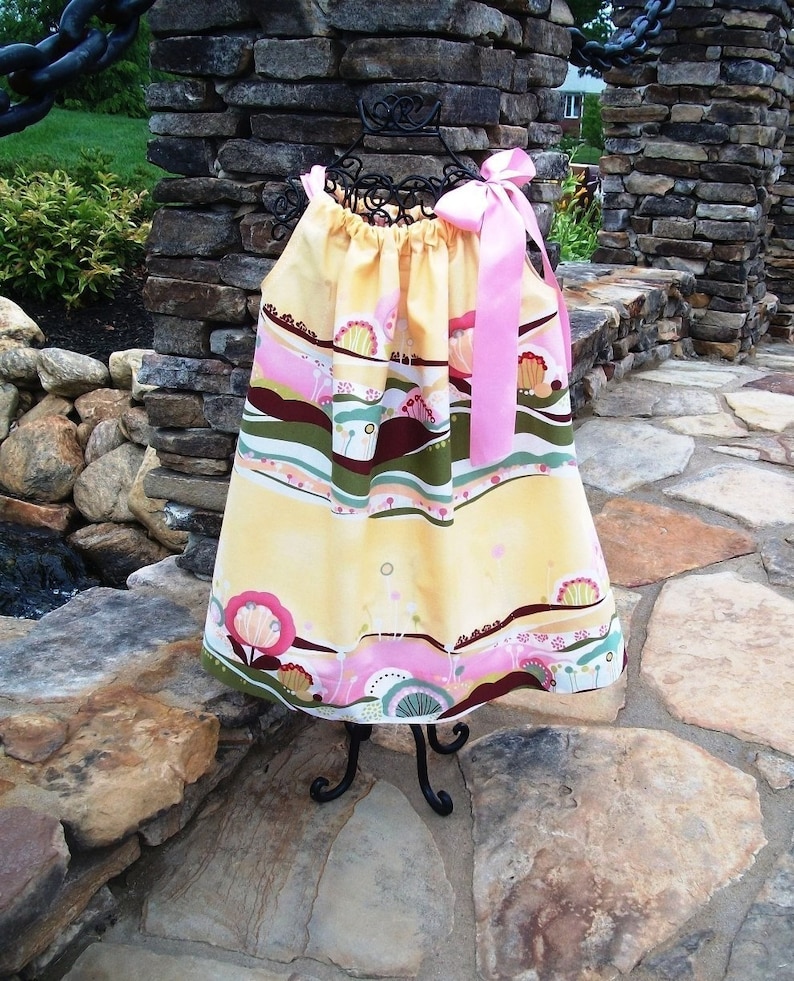 May include: A yellow and pink dress with a floral and landscape print. The dress has a pink ribbon tied around the neck.