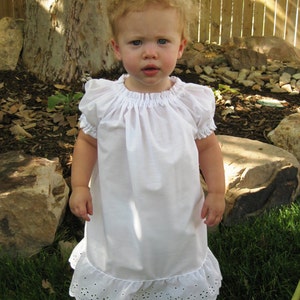 May include: A young child wearing a white dress with short, ruffled sleeves and a ruffled hem. The dress has a gathered neckline. The child is standing on green grass in front of a tree and a wooden fence.