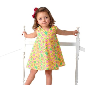 May include: A young girl wearing a yellow sleeveless dress with a pink and green floral pattern. She is standing on a white wooden platform and holding onto a white railing.