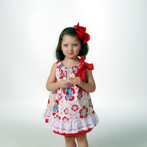 May include: A young girl wearing a white and red floral dress with a white lace trim. She is wearing a red bow in her hair and has a sweet expression on her face.