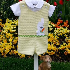 May include: A yellow and white baby romper with a green trim and a white bunny with a carrot applique. The romper is hanging on a white stand against a background of yellow flowers.
