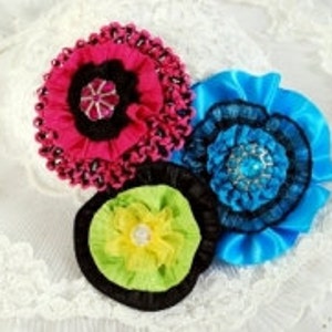 Puede incluir: Tres broches de flores de tela en rosa, azul y amarillo con detalles negros. Cada broche tiene un adorno central con un strass.