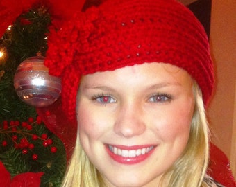 May include: A red crocheted hat with a flower detail. The hat is worn by a person with blonde hair and a bright smile.