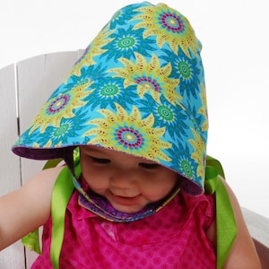 May include: A baby wearing a blue, green, and yellow floral print bonnet with green ribbon ties. The bonnet is reversible with a purple lining. The baby is wearing a pink dress.