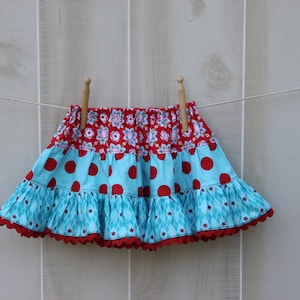 May include: A multi-layered skirt with a red, white, and blue floral print on the top layer, red polka dots on the middle layer, and a blue and white geometric print on the bottom layer. The skirt has a scalloped hem and is hanging on a clothesline.