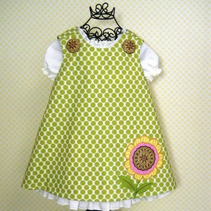 May include: A green sleeveless dress with white polka dots and a large pink and yellow flower applique. The dress has a white ruffle at the bottom and two brown buttons on the shoulders.