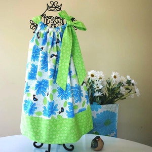 May include: A green and blue floral print dress with a green bow. The dress is on a black stand and a white table. There is a small bird figurine on the table.
