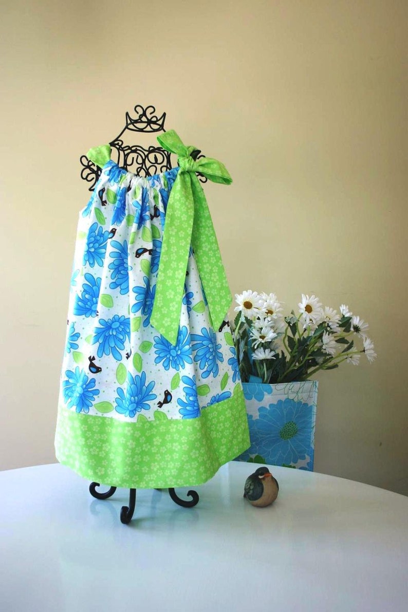May include: A green and blue floral print dress with a green bow. The dress is on a black stand and a white table. There is a small bird figurine on the table.
