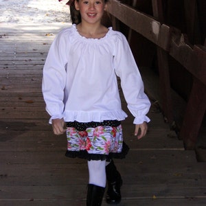 May include: A young girl wearing a white blouse with a ruffled hem, a floral print skirt, white tights, and black boots. She is standing on a wooden bridge.