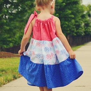 Puede incluir: Una ni&ntilde;a peque&ntilde;a con un vestido rosa, blanco y azul con un estampado de fuegos artificiales. El vestido tiene un dobladillo con volantes y un lazo en el hombro.