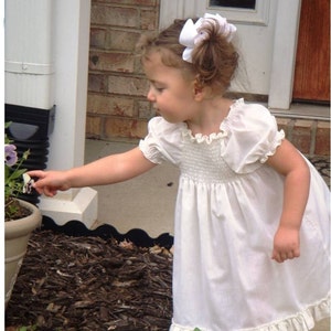 May include: A young girl in a white smocked dress with a lace trim, standing in a garden setting. She is wearing a white bow in her hair and is reaching for a flower.