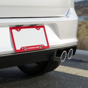 Peut inclure: Un cadre de plaque d'immatriculation rouge avec le texte "C.D. GUADALAJARA" et deux logos. Le cadre est fixé à l'arrière d'une voiture blanche.