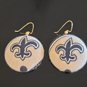 May include: A pair of gold hoop earrings with a round fabric charm featuring the New Orleans Saints logo. The charms are beige with black fleur-de-lis designs.