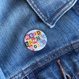 Może przedstawiać: Okrągła przypinka z napisem "READ BANNED BOOKS" w kolorowych literach jest przypięta do niebieskiej kurtki dżinsowej. Projekt przypinki zawiera kolaż różnych kolorów, a kurtka ma brązowe szwy.