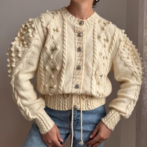 Puede incluir: Un cárdigan de punto color crema con patrones de trenzas, mangas abullonadas con detalles de pompones y motivos florales bordados. La prenda tiene cuello redondo, dobladillo acanalado con cordón ajustable y botones plateados al frente.
