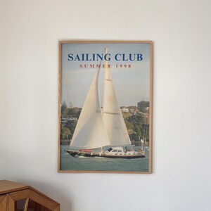 Peut inclure: Une affiche murale encadrée représentant un voilier sur l'eau avec des voiles blanches et une coque sombre. Le texte SAILING CLUB SUMMER 1998 est imprimé en bleu et rouge en haut. L'affiche est dans un cadre en bois clair avec une vitre transparente.