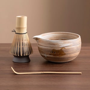 Puede incluir: Un cuenco de matcha de cerámica marrón y blanco con un diseño de gato, un batidor de matcha de bambú y una cuchara de matcha de bambú sobre una superficie de madera.