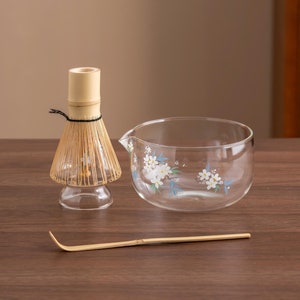 Puede incluir: Un conjunto de tres herramientas para la ceremonia del té japonesa: un batidor de bambú, una jarra de vidrio con un diseño floral y una cuchara de té de bambú. Las herramientas están dispuestas sobre una mesa de madera.