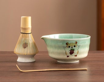 May include: A green and white ceramic matcha bowl with a cartoon hamster design. The bowl is next to a bamboo matcha whisk and a bamboo whisk holder.