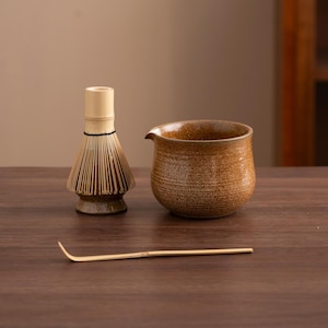 Puede incluir: Un cuenco de matcha de cerámica marrón, un batidor de matcha de bambú y una cuchara de matcha de bambú sobre una superficie de madera.