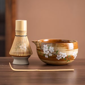 Puede incluir: Un juego de té matcha con un batidor de bambú, un cuenco de cerámica con boquilla y una cuchara de bambú. El cuenco y el batidor tienen diseños florales blancos. El juego está sobre una superficie de madera.