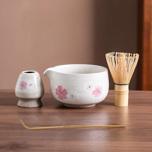 Puede incluir: Un juego de té matcha de cerámica blanca con diseños florales rosas. El juego incluye un cuenco, un batidor, una cuchara y un recipiente pequeño. El batidor es de bambú. Los artículos están dispuestos sobre una superficie de madera.