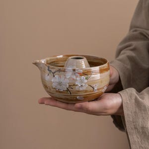 May include: A ceramic teapot with a brown and white glaze and a floral design. The teapot has a round body and a spout on the side. The teapot is decorated with a pattern of white flowers and brown lines.