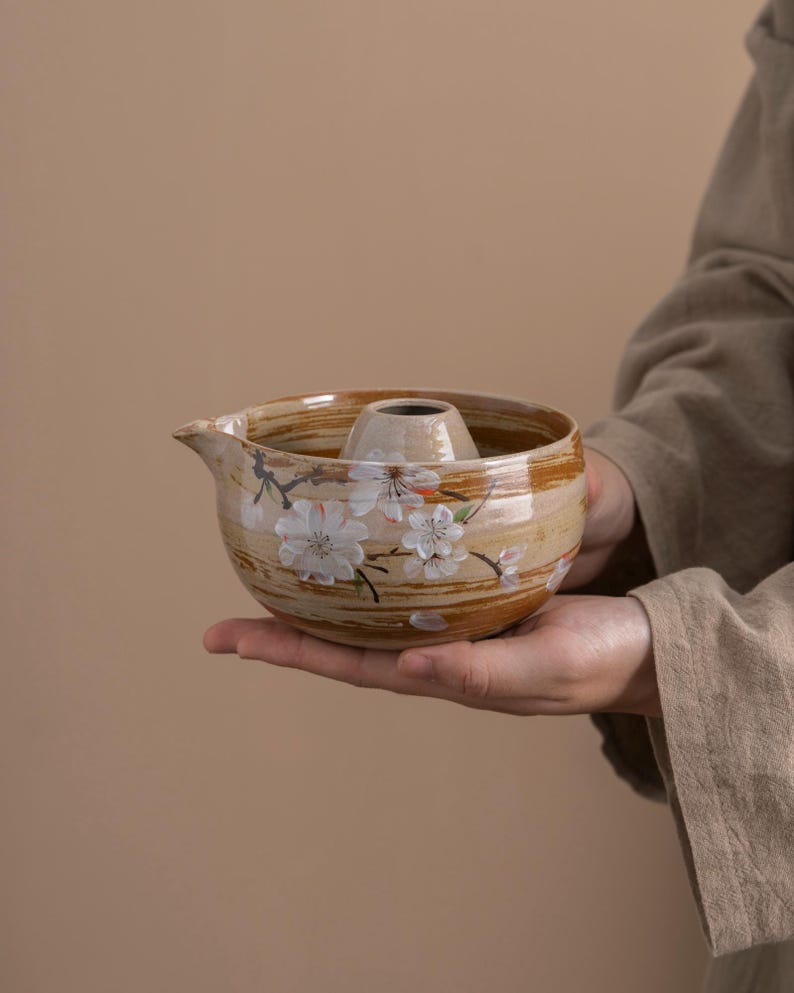 May include: A ceramic teapot with a brown and white glaze and a floral design. The teapot has a round body and a spout on the side. The teapot is decorated with a pattern of white flowers and brown lines.
