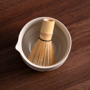 May include: A ceramic matcha bowl with a spout, holding a bamboo matcha whisk. The bowl is off-white with speckled detailing. The whisk is made of thin, light-colored bamboo tines bound together with a black thread. The background is a dark wood surface.