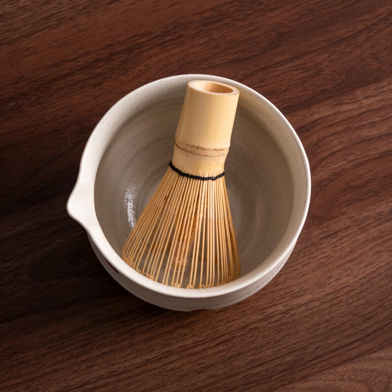 May include: A ceramic matcha bowl with a spout, holding a bamboo matcha whisk. The bowl is off-white with speckled detailing. The whisk is made of thin, light-colored bamboo tines bound together with a black thread. The background is a dark wood surface.