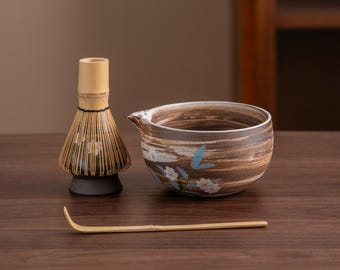 Op de afbeelding: Een bruine en witte keramische matcha-kom met een bloemenpatroon, een bamboe matcha-garde en een bamboe matcha-schep op een houten oppervlak.