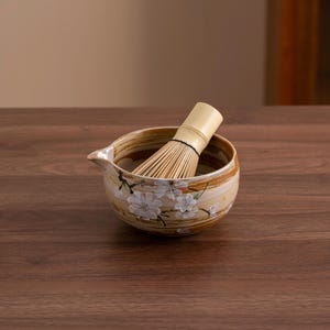 May include: A brown ceramic matcha bowl with a white floral design and a bamboo whisk inside. The bowl is sitting on a wooden surface.