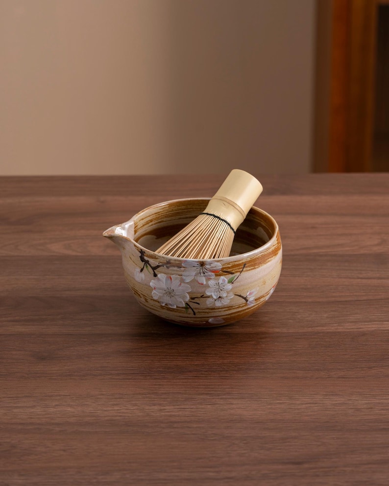 May include: A brown ceramic matcha bowl with a white floral design and a bamboo whisk inside. The bowl is sitting on a wooden surface.