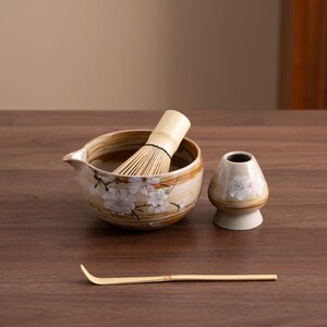 May include: A brown and white ceramic matcha bowl with a floral design, a bamboo whisk, a small ceramic cup with a floral design, and a bamboo matcha scoop.