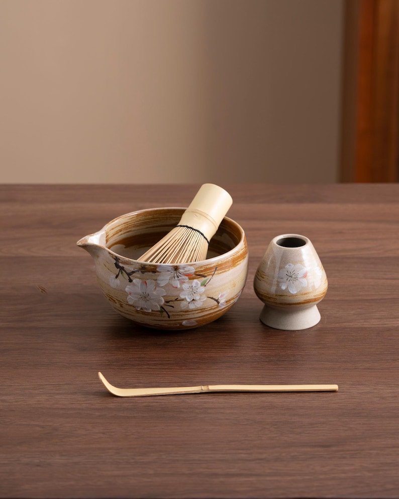 May include: A brown and white ceramic matcha bowl with a floral design, a bamboo whisk, a small ceramic cup with a floral design, and a bamboo matcha scoop.