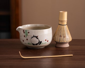 Op de afbeelding: Een witte keramische matcha-kom met een ontwerp van een zwarte kat en rode kersen, een bamboe garde en een bamboe matcha-lepel.