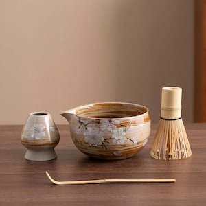 May include: A brown and white ceramic tea bowl with a floral design, a small brown and white ceramic tea scoop, a bamboo whisk, and a bamboo whisk handle.