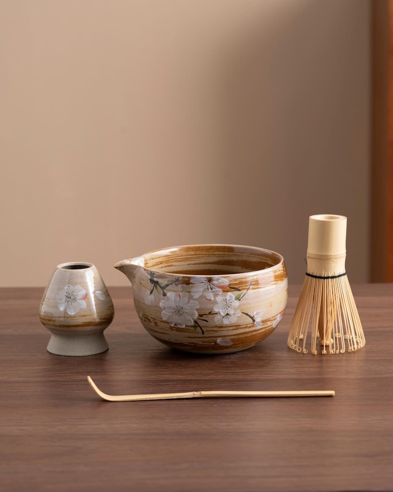 May include: A brown and white ceramic tea bowl with a floral design, a small brown and white ceramic tea scoop, a bamboo whisk, and a bamboo whisk handle.