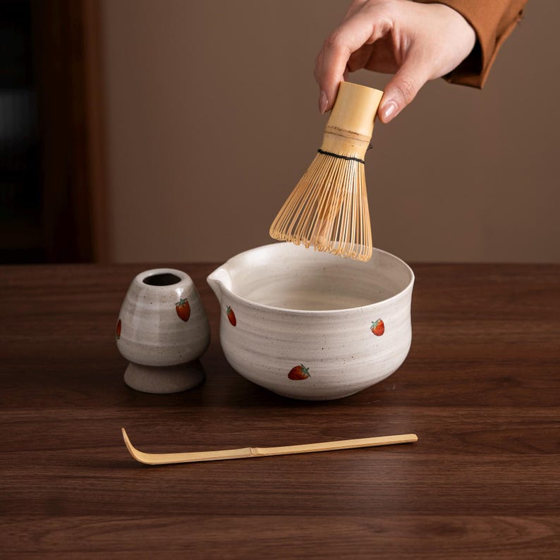 May include: A matcha tea set with a bamboo whisk, a ceramic bowl, a small ceramic container, and a bamboo scoop. The bowl and container are white with red strawberry designs. The whisk is held above the bowl.